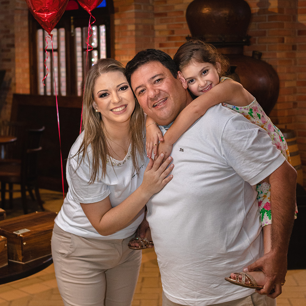 Foto de Andressa, Ricardo e Júlia. Eles estão lhando para a câmera, sorrindo, e a Júlia está nas costas do Ricardo, segurando em seus ombros