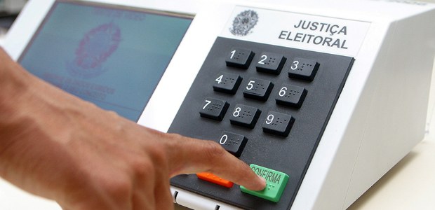 A imagem mostra uma urna eletrônica, ao lado direito os botões numéricos e acima as palavras: Justiça Eleitoral. Aparece uma mão de um eleitor prestes a apertar o botão.